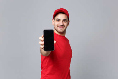 Delivery man in red uniform hold pc cellphone isolated on grey wall background studio portrait. Professional fun male employee in cap t-shirt print working courier. Service concept. Mock up copy spaceの写真素材