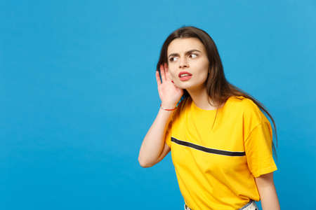 Portrait of curious young woman in vivid casual clothes looking aside, try to hear you with hand...