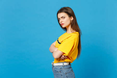 Side view of displeased dissatisfied young woman in vivid casual clothes looking camera, holding hands crossed isolated on blue wall background in studio. People lifestyle concept. Mock up copy space.の写真素材