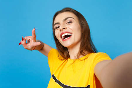 Close up selfie shot of cheerful cute young woman in vivid casual clothes looking camera, showing victory sign isolated on blue wall background in studio. People lifestyle concept. Mock up copy space.の写真素材