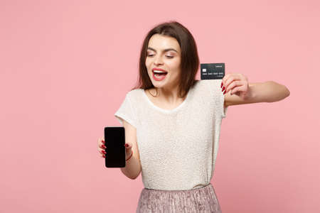 Pretty young woman in casual light clothes posing isolated on pastel pink wall background. People lifestyle concept. Mock up copy space. Holding mobile phone with blank empty screen, credit bank card.の写真素材