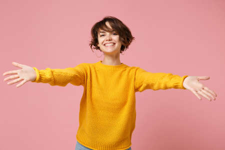 Smiling young brunette woman girl in yellow sweater posing isolated on pastel pink wall background studio portrait. People lifestyle concept. Mock up copy space. Standing with outstretched hands.の写真素材
