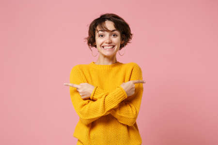 Smiling young brunette woman girl in yellow sweater posing isolated on pastel pink background in studio. People lifestyle concept. Mock up copy space. Point index fingers aside, holding hands crossed.の写真素材