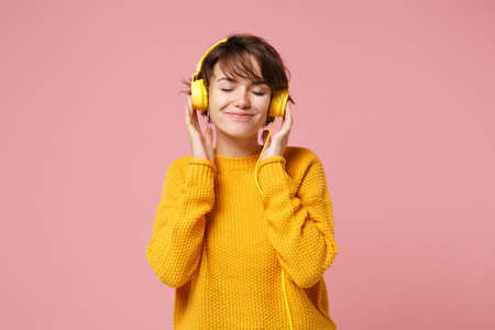 Relaxed young brunette woman girl in yellow sweater posing isolated on pastel pink background in studio. People lifestyle concept. Mock up copy space. Listen music with headphones keeping eyes closed.の写真素材