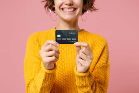 Cropped image of smiling young brunette woman girl in yellow sweater posing isolated on pastel pink wall background studio portait. People lifestyle concept. Mock up copy space. Hold credit bank card.の写真素材
