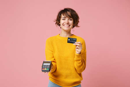 Funny young brunette woman in yellow sweater posing isolated on pink background. People lifestyle concept. Mock up copy space. Hold bank payment terminal to process and acquire credit card payments.の写真素材