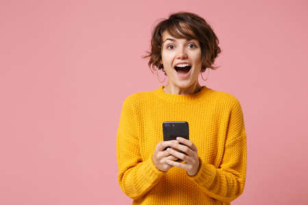 Excited young brunette woman girl in yellow sweater posing isolated on pastel pink wall background studio portait. People lifestyle concept. Mock up copy space. Using mobile phone, typing sms message.の写真素材