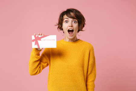 Shocked young brunette woman girl in yellow sweater posing isolated on pastel pink background studio portrait. People sincere emotions lifestyle concept. Mock up copy space. Holding gift certificate.の写真素材