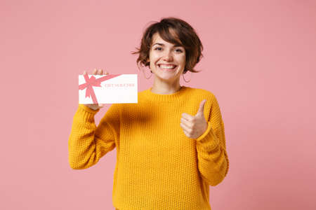 Cheerful young brunette woman girl in yellow sweater posing isolated on pastel pink background studio portrait. People lifestyle concept. Mock up copy space. Holding gift certificate showing thumb up.の写真素材