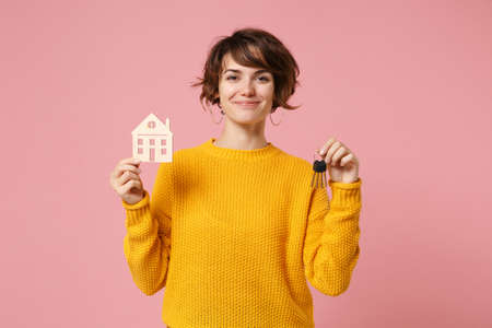 Smiling young brunette woman girl in yellow sweater posing isolated on pastel pink wall background studio portrait. People lifestyle concept. Mock up copy space. Hold in hands house and bunch of keys.の写真素材