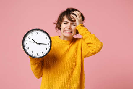 Preoccupied young brunette woman girl in yellow sweater posing isolated on pastel pink wall background. People sincere emotions lifestyle concept. Mock up copy space. Holding clock, put hand on head.の写真素材