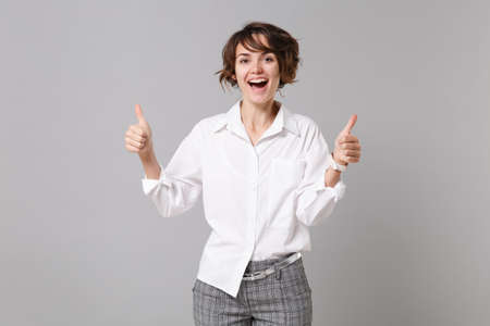 Cheerful funny young brunette business woman in white shirt posing isolated on gray wall background studio portrait. Achievement career wealth business concept. Mock up copy space. Showing thumbs upの写真素材