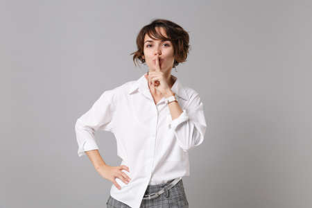 Secret young business woman in white shirt posing isolated on gray wall background. Achievement career wealth business concept. Mock up copy space. Say hush be quiet with finger on lips shhh gestureの写真素材