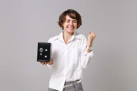 Joyful young business woman in white shirt isolated on gray background. Achievement career wealth business concept. Mock up copy space Doing winner gesture hold metal bank safe for money accumulationの写真素材