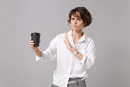 Displeased young business woman in white shirt isolated on gray background. Achievement career wealth business concept. Mock up copy space Showing stop gesture with palm to paper cup of coffee or teaの写真素材