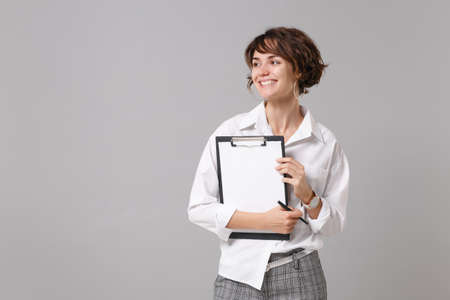 Cheerful young business woman in white shirt posing isolated on gray wall background. Achievement career wealth business concept. Mock up copy space. Hold clipboard with blank empty sheet workspaceの写真素材