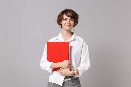 Smiling young business woman in white shirt posing isolated on gray background studio portrait. Achievement career wealth business concept. Mock up copy space. Holding red folder for papers documentの写真素材