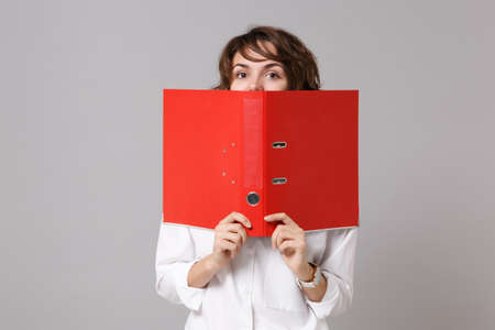 Funny young business woman in white shirt posing isolated on gray background. Achievement career wealth business concept. Mock up copy space. Hiding, covering face with red folder for papers documentの写真素材