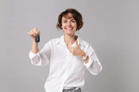 Funny young business woman in white shirt posing isolated on gray wall background studio portrait. Achievement career wealth business concept. Mock up copy space. Showing thumb up, holding car keysの写真素材