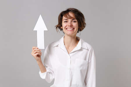 Cheerful funny young business woman in white shirt posing isolated on gray wall background studio portrait. Achievement career wealth business concept. Mock up copy space. Holding in hand up arrowの写真素材