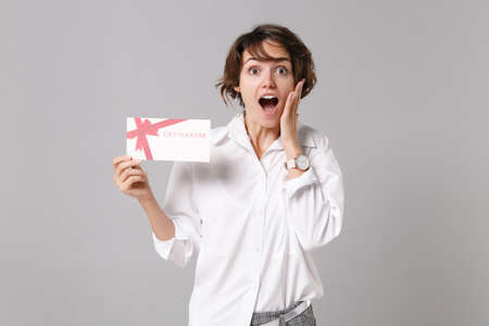 Shocked young business woman in white shirt posing isolated on grey background studio portrait. Achievement career wealth business concept. Mock up copy space. Hold gift certificate put hand on cheek.の写真素材