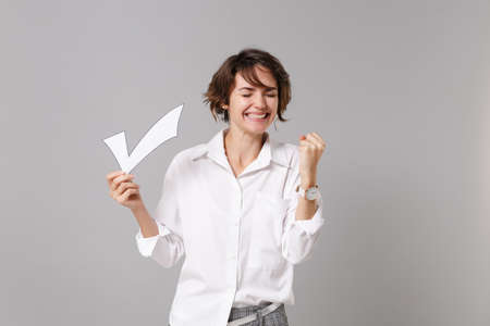 Happy young business woman in white shirt posing isolated on grey wall background in studio. Achievement career wealth business concept. Mock up copy space. Hold paper check mark doing winner gesture.の写真素材