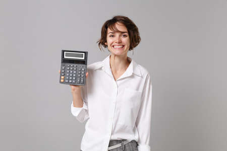 Cheerful pretty young business woman in white shirt posing isolated on grey wall background studio portrait. Achievement career wealth business concept. Mock up copy space. Hold in hand calculator.の写真素材