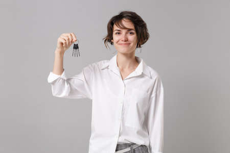 Smiling beautiful young business woman in white shirt posing isolated on grey wall background studio portrait. Achievement career wealth business concept. Mock up copy space. Holding bunch of keys.の写真素材