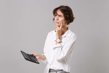 Pensive young business woman in white shirt posing isolated on grey wall background in studio. Achievement career wealth business concept. Mock up copy space. Hold calculator put hand prop up on chin.の写真素材