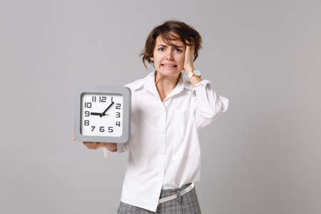 Preoccupied young business woman in white shirt posing isolated on grey wall background studio portrait. Achievement career wealth business concept. Mock up copy space. Hold clock, put hand on head.の写真素材