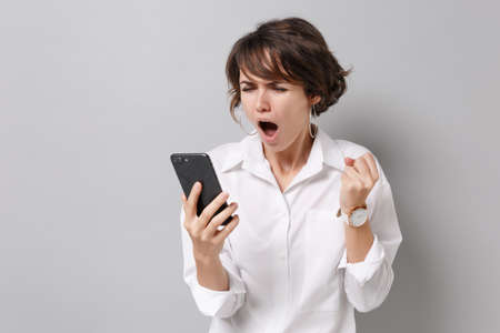 Angry irritated young business woman in white shirt posing isolated on grey background studio portrait. Achievement career wealth business concept. Mock up copy space. Holding mobile phone, swearing.の写真素材