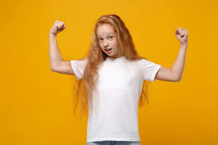 Strong little ginger kid girl 12-13 years old in white t-shirt isolated on bright yellow background children studio portrait. Childhood lifestyle concept. Mock up copy space. Showing biceps muscles.の写真素材