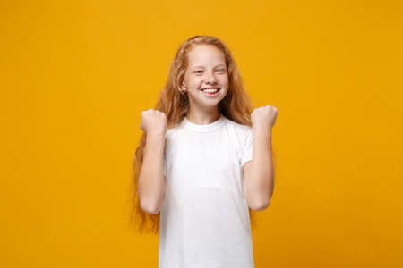 Smiling little ginger kid girl 12-13 years old in white t-shirt isolated on bright yellow background children studio portrait. Childhood lifestyle concept. Mock up copy space. Doing winner gesture.の写真素材