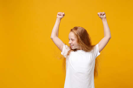 Little ginger kid girl 12-13 years old in white t-shirt isolated on yellow wall background children portrait. Childhood lifestyle concept. Mock up copy space. Rising hands clenching fists like winner.の写真素材