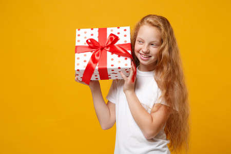 Cute little ginger kid girl 12-13 years old in white t-shirt isolated on yellow background. New Year Womens Day birthday, holiday concept. Mock up copy space. Hold present box with gift ribbon bow.の写真素材