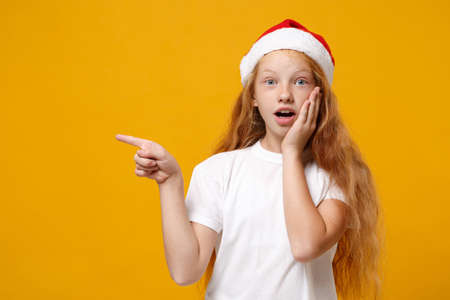 Shocked little ginger kid Santa girl 12-13 years old in white t-shirt Christmas hat isolated on yellow background. New Year 2020 celebration concept. Mock up copy space. Pointing index finger aside.の写真素材