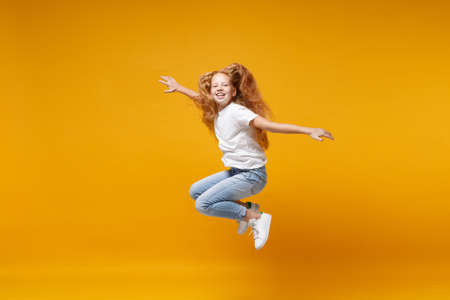 Pretty little ginger kid girl 12-13 years old in white t-shirt isolated on yellow background. Childhood lifestyle concept. Mock up copy space. Having fun, fooling around, jumping, spreading hands.の写真素材