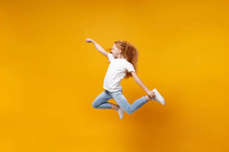 Side view of little ginger kid girl 12-13 years old in white t-shirt isolated on yellow background children portrait. Childhood lifestyle concept. Mock up copy space. Having fun, fooling around, jump.の写真素材