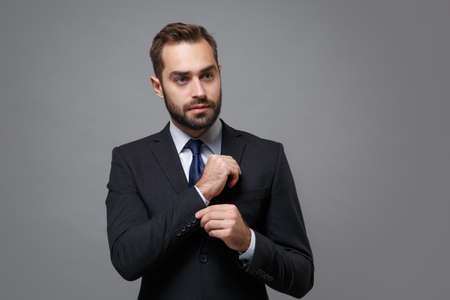 Confident young business man in classic black suit shirt tie posing isolated on grey background. Achievement career wealth business concept. Mock up copy space. Straightening sleeves, looking aside.の写真素材