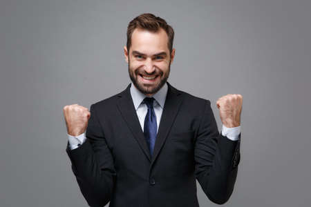 Smiling young business man in classic black suit shirt tie posing isolated on grey background in studio. Achievement career wealth business concept. Mock up copy space. Clenching fists like winner.の写真素材