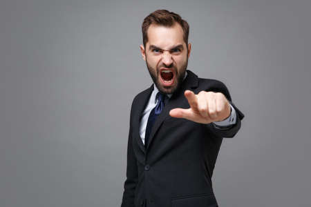 Angry young bearded business man in suit shirt tie posing isolated on grey background. Achievement career wealth business concept. Mock up copy space. Point index finger on camera, screaming swearing.の写真素材