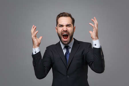 Angry mad young business man in classic black suit shirt tie posing isolated on grey background. Achievement career wealth business concept. Mock up copy space. Spreading hands, screaming swearing.の写真素材