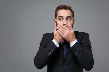 Shocked young bearded business man in classic black suit shirt tie posing isolated on grey wall background. Achievement career wealth business concept. Mock up copy space. Covering mouth with hands.の写真素材
