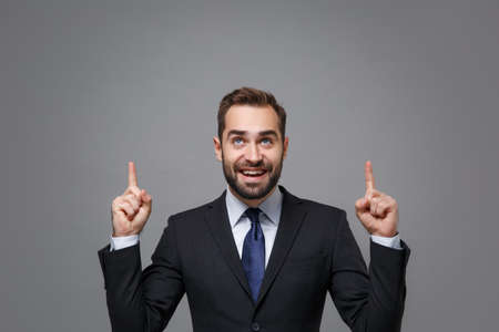 Handsome young bearded business man in classic black suit shirt tie posing isolated on grey wall background. Achievement career wealth business concept. Mock up copy space. Pointing index fingers up.の写真素材