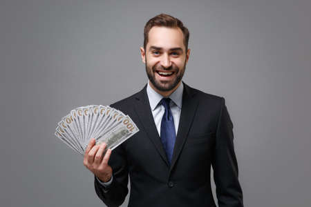 Cheerful young business man in classic suit shirt tie posing isolated on grey background. Achievement career wealth business concept. Mock up copy space. Holding fan of cash money in dollar banknotes.の写真素材