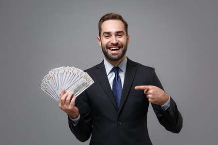 Laughing young business man in suit shirt tie posing isolated on grey background. Achievement career wealth business concept. Mock up copy space. Point finger on fan of cash money in dollar banknotes.の写真素材