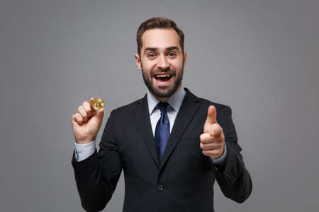 Excited young business man in suit shirt tie posing isolated on grey background. Achievement career wealth business concept. Mock up copy space. Hold bitcoin future currency pointing finger on camera.の写真素材