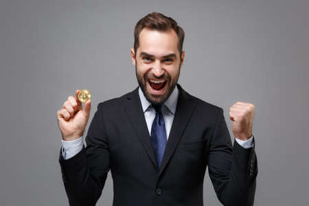 Joyful young business man in black suit shirt tie posing isolated on grey background. Achievement career wealth business concept. Mock up copy space. Hold bitcoin future currency doing winner gesture.の写真素材