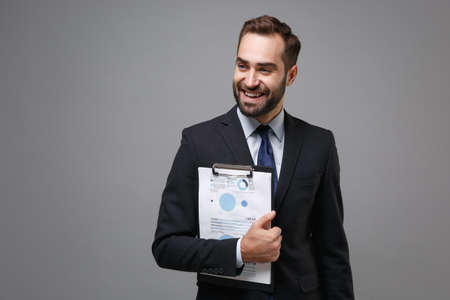 Cheerful young business man in suit shirt tie posing isolated on grey background. Achievement career wealth business concept. Mock up copy space. Hold clipboard with papers document, looking aside.の写真素材