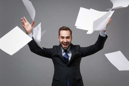 Laughing cheerful young business man in classic black suit shirt tie posing isolated on grey wall background. Achievement career wealth business concept. Mock up copy space. Throwing work documents.の写真素材